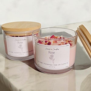May include: Two glass candle jars with wooden lids. The jars are filled with a pink candle and rose petals. The labels on the jars read "Corners & Candles Rose Soy Candle | Aromatherapy | Made with Essential Oils 8 oz (227 g)"