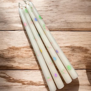 Może przedstawiać: Cztery białe świece stożkowe z pastelowym wzorem jajek wielkanocnych i marchewek.