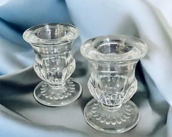 May include: Two clear glass candle holders featuring a wide, flared rim, a rounded body with vertical ribbed detailing, and a circular base with a scalloped edge pattern. The glass is transparent and smooth, designed to hold a single taper candle.