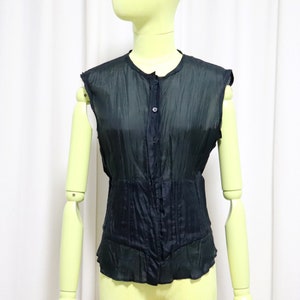 Pode incluir: Top preto sem mangas com botões na frente e cintura ajustada. O top tem um tecido texturizado e semitransparente e um decote redondo. A peça de roupa é exibida num manequim.