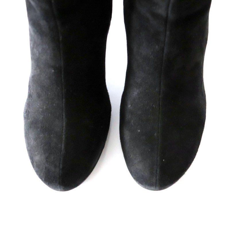 May include: A pair of black suede boots, viewed from the toe up. The boots have a soft, matte texture and a classic design. The image is set against a clean white background.