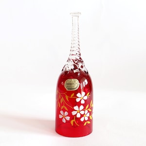 Peut inclure: Un vase en verre rouge avec un col torsadé transparent et une base évasée. Le vase est décoré de fleurs blanches et de feuilles dorées. Une petite étiquette est apposée sur le devant du vase.