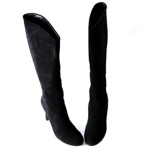 May include: A pair of black knee-high boots. The boots are made of a soft, suede-like material. They have a pointed toe and a small, curved heel. The boots are set against a white background.