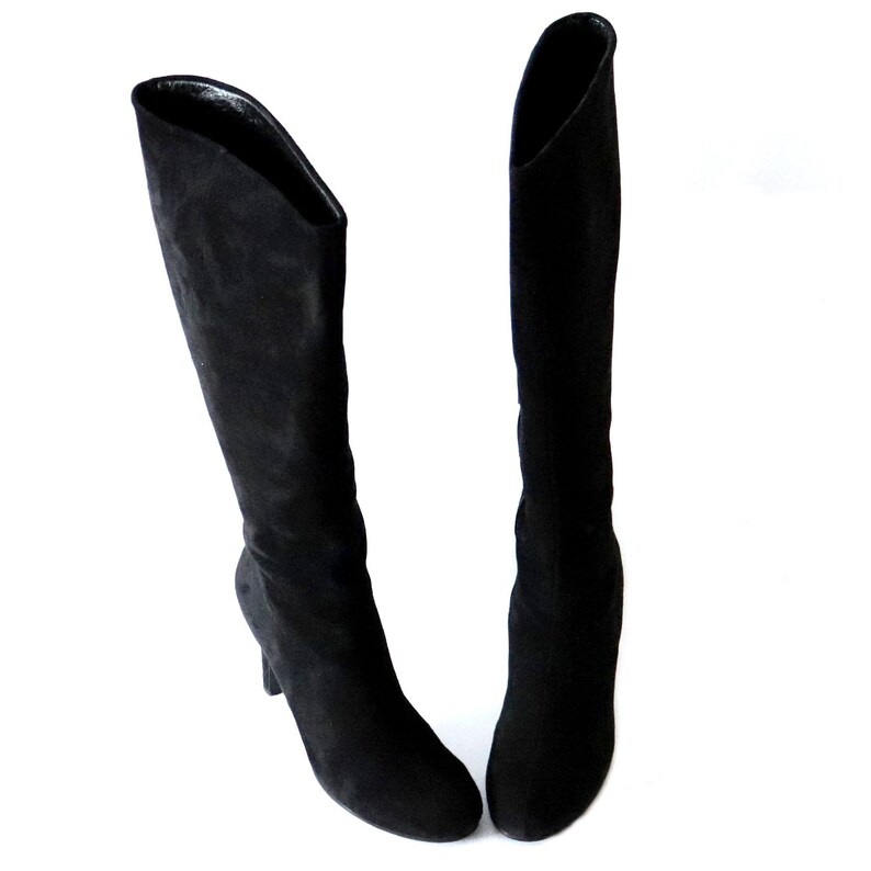 May include: A pair of black knee-high boots. The boots are made of a soft, suede-like material. They have a pointed toe and a small, curved heel. The boots are set against a white background.