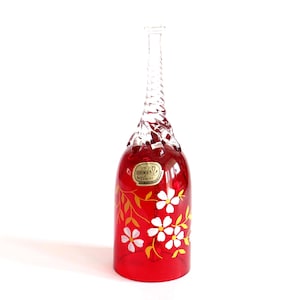 Könnte beinhalten: Eine rote böhmische Glasvase mit einem klaren, gedrehten Hals und einem ausgestellten Boden. Die Vase ist mit weißen Blumen und goldenen Blättern verziert. Ein kleines Etikett ist auf der Vorderseite angebracht. Die Vase ist ca. 25 cm hoch.