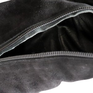 May include: Close-up of a black suede bag with a partially open zipper. The interior of the bag is visible, revealing a dark leather lining. The zipper is silver and runs along the top edge of the bag.