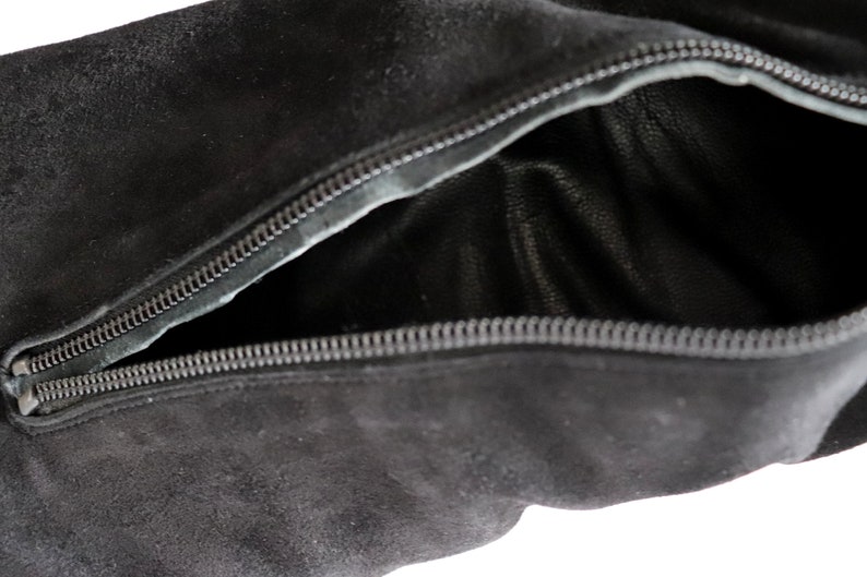 May include: Close-up of a black suede bag with a partially open zipper. The interior of the bag is visible, revealing a dark leather lining. The zipper is silver and runs along the top edge of the bag.