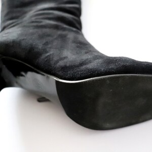 May include: Close-up of a black suede boot with a high heel. The boot's sole and heel are glossy black, contrasting with the matte texture of the suede upper. The heel is approximately 3 inches high.