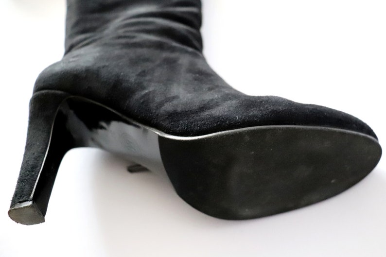 May include: Close-up of a black suede boot with a high heel. The boot's sole and heel are glossy black, contrasting with the matte texture of the suede upper. The heel is approximately 3 inches high.