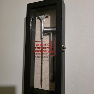Puede incluir: Un expositor enmarcado en negro con frente de cristal, que contiene un martillo de emergencia plateado y una herramienta para romper cristales. El texto "IN CASE OF EMERGENCY BREAK GLASS" está impreso en rojo. El fondo es de tela beige.