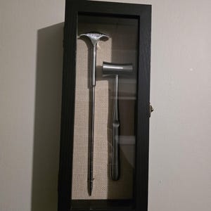 Puede incluir: Una exhibición enmarcada con dos instrumentos médicos antiguos. Los instrumentos son plateados, uno con un mango largo y delgado y una cabeza ensanchada, el otro una pequeña herramienta en forma de martillo. El marco es negro con un respaldo de tela neutra.