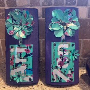 May include: Two handmade wooden wall art pieces. Each features a purple painted base with a turquoise and pink floral design. The flowers are made from recycled materials. One piece has the text "Zon" and "V/cts". The other has the text "AN AMERICAN".