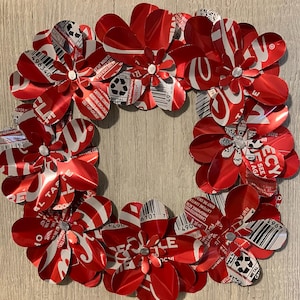 May include: A handcrafted wreath made from recycled red and white aluminum can flowers. The flowers are arranged in a circular pattern, with silver accents in the center of each flower. The wreath is a unique, eco-friendly decoration.