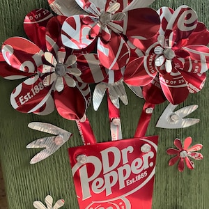 May include: A handcrafted floral arrangement made from recycled Dr. Pepper cans. The bouquet features red and white flowers with silver accents, arranged in a Dr. Pepper can vase, mounted on a green wooden background.