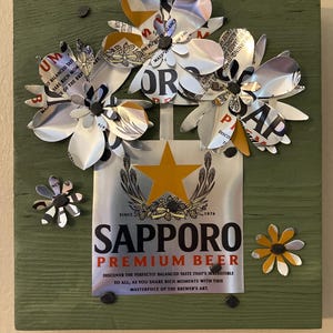 May include: A green wooden sign with a Sapporo Premium Beer can attached to it. The can has a gold star and the text "Since 1976".  The can is surrounded by flowers made from aluminum cans.