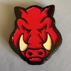 May include: A decorative emblem shaped like a stylized boar head, featuring a vibrant red face with black outlines and textured black ears. The design includes two prominent cream-colored tusks and a red snout, all rendered in a raised, layered relief.