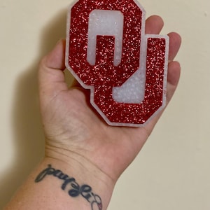 May include: A red glittery "OU" emblem with a white interior, held in a hand. The letters are outlined in red glitter, with a white interior. The emblem is a decorative item, possibly for a fan or collector.