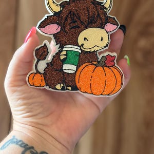 May include: A glittery, die-cut acrylic or resin charm featuring a brown Highland cow with tan horns, holding a green and white coffee cup. The cow sits next to a bright orange pumpkin with a small red leaf on top and a tiny orange pumpkin.