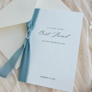 Pode incluir: Um cartão branco com o texto "A Letter to My Best Friend On Her Wedding Day" e a data de 23 de setembro de 2025, está amarrado com uma fita azul e colocado dentro de um envelope correspondente. O cartão e o envelope estão sobre um tecido rosa claro.