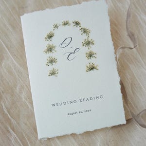 May include: Ivory-colored wedding reading card with a floral arch design. The card features the initials "D & E" and the text "WEDDING READING" and "August 22, 2026". The card has a deckled edge and is set against a neutral fabric background.