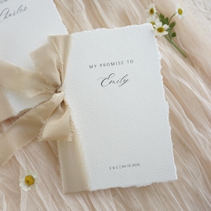 Può includere: Biglietti di promessa di matrimonio color crema con bordi strappati, legati con un nastro beige. Il testo "MY PROMISE TO Emily" è stampato sulla carta con una scrittura elegante. Un rametto di fiori bianchi con centri gialli aggiunge un tocco delicato.