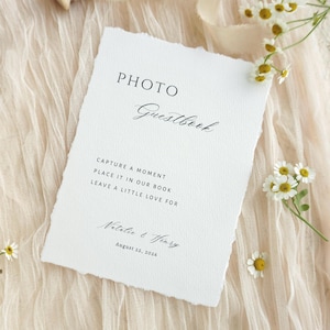 May include: A white Photo Guestbook card with torn edges, featuring elegant script. The card includes the text "Capture a moment, place it in our book, leave a little love for Natalie & Henry, August 22, 2026." The card is on a cream-colored fabric with daisies.
