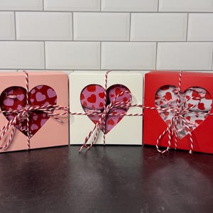 May include: Three heart-shaped gift boxes in pink, white, and red. Each box has a heart-shaped cutout revealing a patterned interior and is tied with red and white twine. The boxes are arranged on a dark surface.