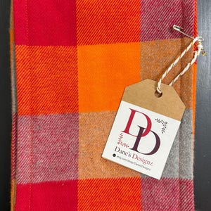 May include: A red, orange, and gray plaid fabric with a brown tag that says "Dane's Designz" and a website address. The fabric has a sewn edge.