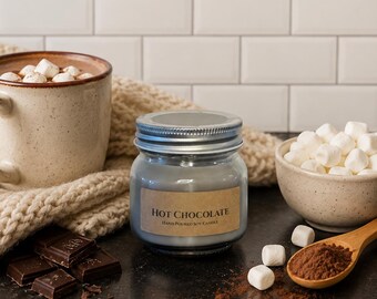 Puede incluir: Una vela en frasco de vidrio con tapa plateada y etiqueta color canela que dice HOT CHOCOLATE HAND POURED SOY CANDLE. El texto Cozy Winter Favorite aparece arriba, y Rich cocoa • sweet cream • fluffy marshmallow aparece debajo de la vela.