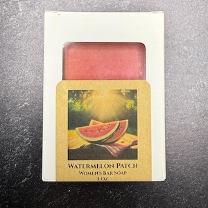 Puede incluir: Una pastilla de jabón rosa en una caja blanca con una etiqueta. La etiqueta presenta una imagen de acuarela de rodajas de sandía y el texto "Watermelon Patch Women's Bar Soap 85g."