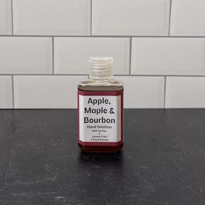 Puede incluir: Una pequeña botella transparente de desinfectante de manos con una etiqueta blanca con el texto "Apple, Maple & Bourbon". La etiqueta también indica que el producto contiene aceite de árbol de té y aceites de vitamina E, y es de 59 ml. La botella tiene una tapa transparente.