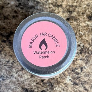 May include: A silver lid with a pink label that reads "Mason Jar Candle" with a flame icon and the text "Watermelon Patch".