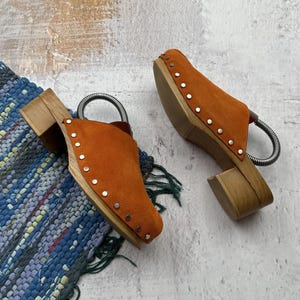 May include: A pair of orange suede clogs with wooden heels and silver studs. The clogs have a slingback strap and are sitting on a blue and white rug.