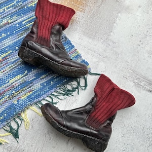 May include: A pair of brown leather boots with red knit cuffs. The boots have a chunky sole and are sitting on a blue and green rug.