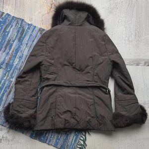 May include: A brown winter coat with a fur-lined hood and cuffs. The coat has a belt at the waist and a quilted design. The coat is displayed on a textured surface with a colorful rug.