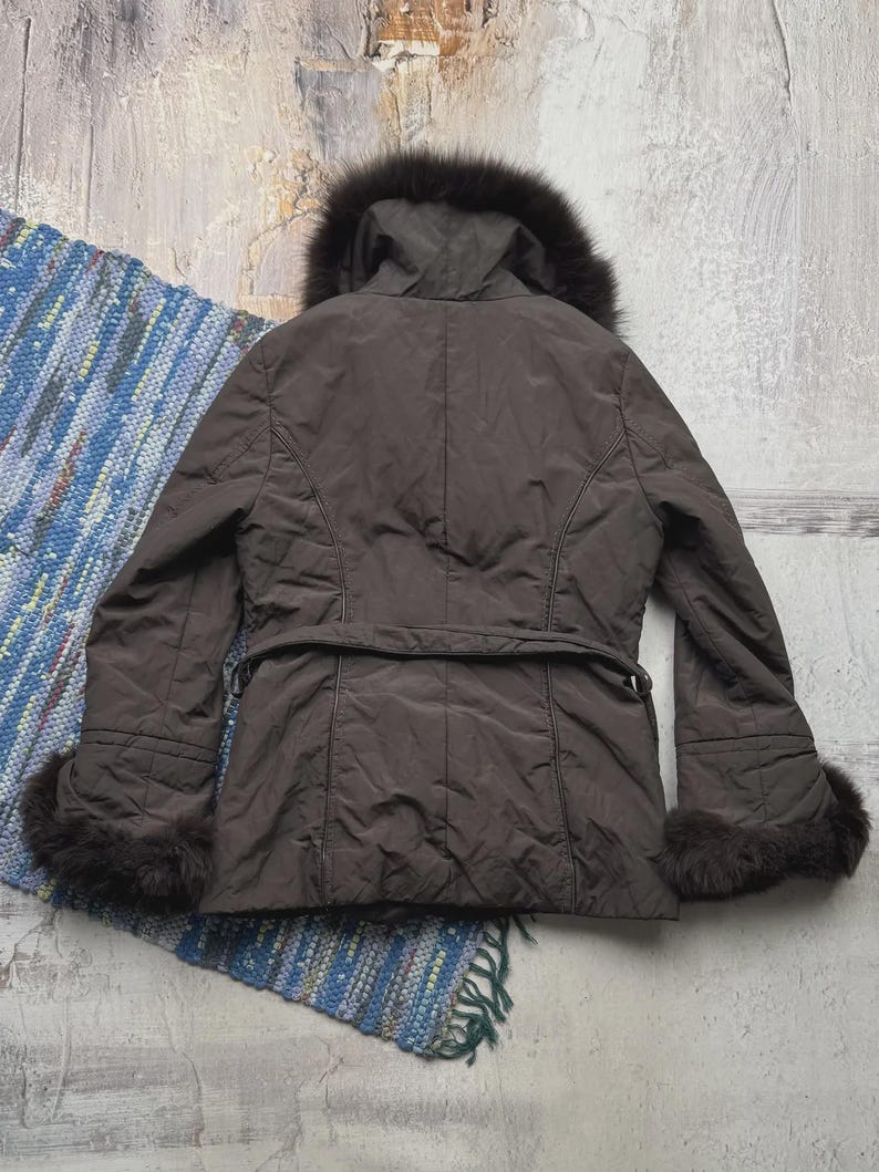 May include: A brown winter coat with a fur-lined hood and cuffs. The coat has a belt at the waist and a quilted design. The coat is displayed on a textured surface with a colorful rug.