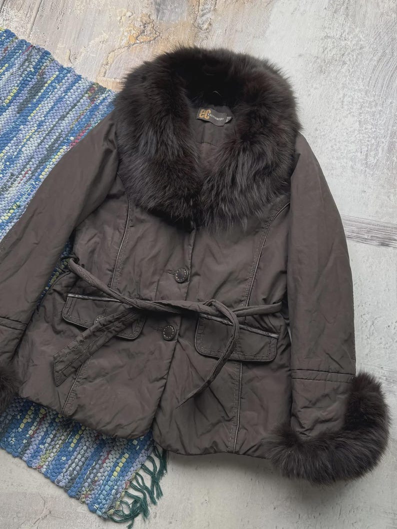 May include: A brown jacket with a fur collar and cuffs. The jacket has a belted waist, two front pockets, and button closures. The fur appears to be dark brown. The jacket is a stylish outerwear piece.