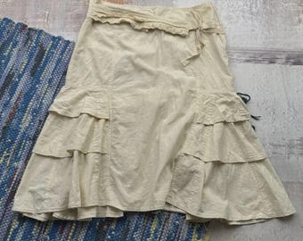 May include: A beige skirt with a tiered, ruffled design. The skirt has a lace detail at the waist and a tie closure. The skirt is made of a lightweight fabric and is suitable for casual wear.