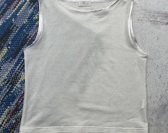May include: A white, sleeveless waffle-knit top with a scoop neck and a small label near the neckline. The top has a textured surface and a satin trim. The background includes a blue and white patterned rug.