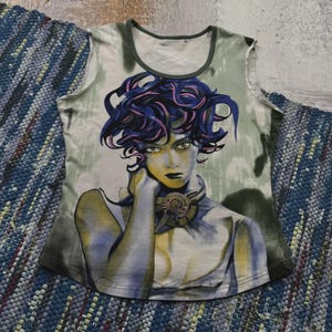 Pode incluir: Camiseta sem mangas com um retrato impresso de uma pessoa com cabelo azul e rosa e uma gola decorativa. A camisa tem decote verde e fundo verde claro e cinza. A camisa é exibida em um tapete tecido azul e cinza.