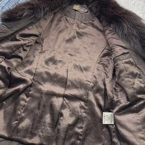 May include: A brown fur-trimmed coat with a satin lining. The coat features a dark brown fur collar and a smooth, shiny interior. The inside of the coat has a label with text. The coat is open, revealing the interior.