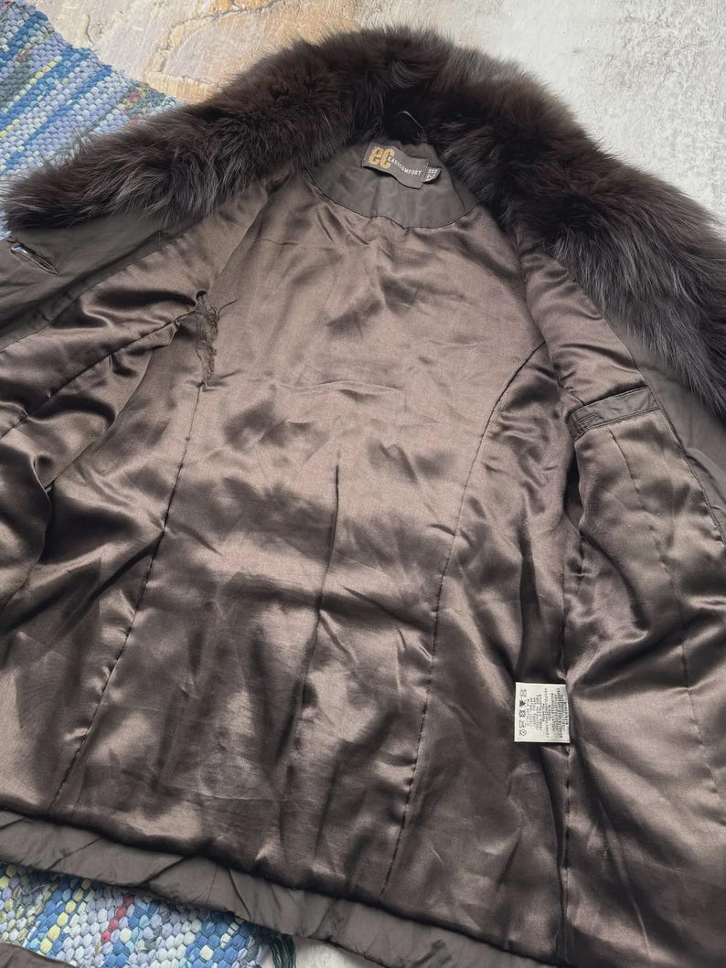 May include: A brown fur-trimmed coat with a satin lining. The coat features a dark brown fur collar and a smooth, shiny interior. The inside of the coat has a label with text. The coat is open, revealing the interior.