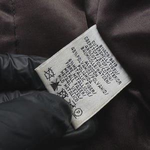 May include: Close-up of a tag with text in multiple languages, including English, French, and German. The tag is white with black text and is sewn into a brown fabric. The tag lists the materials of the garment, including cotton, polyamide, and metallic fiber.