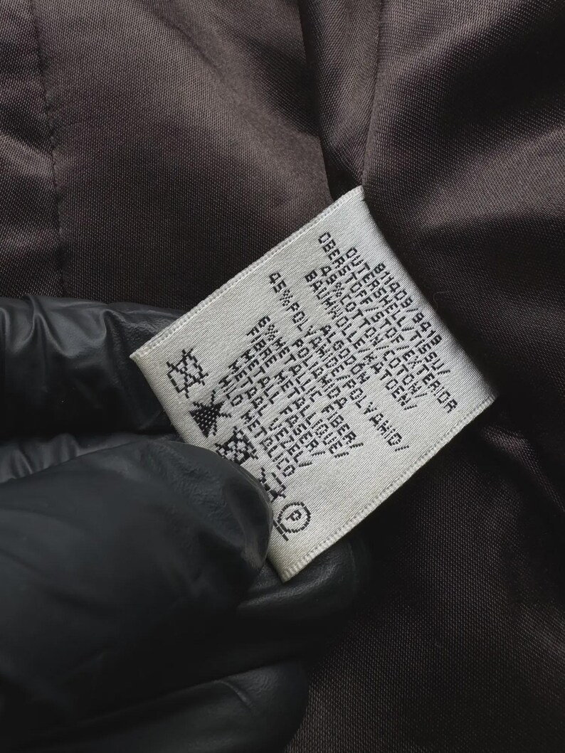 May include: Close-up of a tag with text in multiple languages, including English, French, and German. The tag is white with black text and is sewn into a brown fabric. The tag lists the materials of the garment, including cotton, polyamide, and metallic fiber.