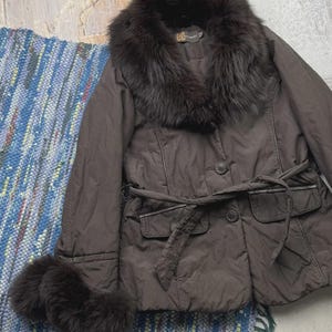 May include: A brown jacket with a fur collar and cuffs. The jacket has a belted waist, two front pockets, and button closures. The jacket is displayed on a blue and white woven rug.