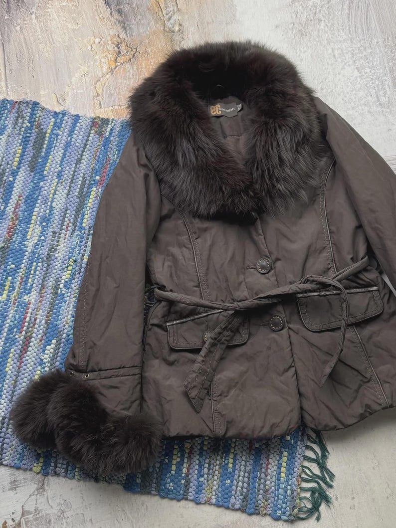 May include: A brown jacket with a fur collar and cuffs. The jacket has a belted waist, two front pockets, and button closures. The jacket is displayed on a blue and white woven rug.