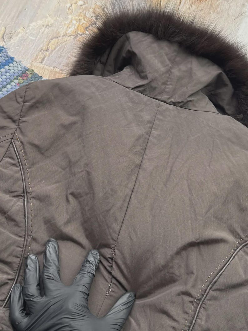 May include: A brown jacket with a hood trimmed with dark fur. The jacket has a quilted design and visible stitching details. The image shows the back of the jacket.