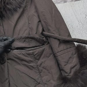 May include: A brown coat with a fur collar and cuffs. The coat has a button closure, a pocket, and a belt. The fur trim is dark brown. The coat appears to be made of a smooth, possibly synthetic material.