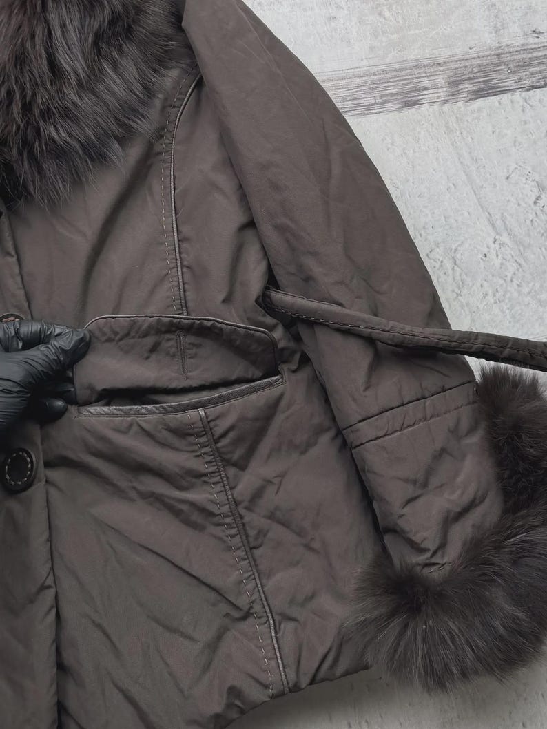 May include: A brown coat with a fur collar and cuffs. The coat has a button closure, a pocket, and a belt. The fur trim is dark brown. The coat appears to be made of a smooth, possibly synthetic material.