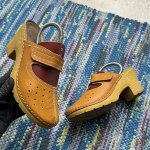May include: A pair of tan and dark red leather clogs featuring a wooden block heel, a perforated toe box, and a hook-and-loop strap closure across the instep. The shoes have a light-colored wooden sole with a textured tread and a metal shoe tree.